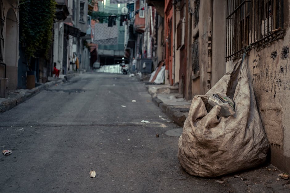 A large, weathered, and dirty beige fabric trash bag sits on the uneven asphalt pavement of a narrow urban street, leaning against a graffitied concrete wall with iron window bars. The bag appears to be filled with miscellaneous waste, with some plastic and paper items partially protruding from the top. The street is lined with closely packed, multi-story residential buildings featuring worn facades, with visible signs of age and some graffiti on the walls. In the background, laundry is hanging across the street, and a slight incline leads towards a distant archway or opening at the end of the street. Scattered debris, including small pieces of paper and rubbish, is seen on the ground around the bag. The environment reflects a typical densely populated urban area where clutter and waste are accumulated on the street, emphasizing the importance of private waste collection services like those offered by Waste Clearance Kensington for on-site rubbish removal and alternative waste handling solutions in such challenging environments.