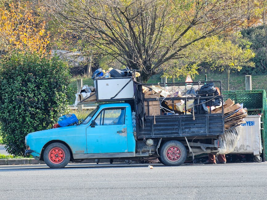 A small, old blue pickup truck parked on the side of a street, heavily loaded with various types of rubbish including cardboard boxes, black plastic bags, wooden planks, and miscellaneous waste materials. The truck's cab is weathered, with faded paint and visible signs of wear, while the flatbed area is filled to capacity, with some items stacked and secured with ropes. The truck is positioned adjacent to a green hedge and a large tree with yellowing leaves, indicating an autumn setting. In the background, a metal fence and a grassy area are visible, suggesting a residential or park environment. The scene is illuminated by natural daylight, casting soft shadows on the pavement. The image relates to private waste collection and rubbish removal services, such as those offered by Waste Clearance Kensington, especially in contexts of alternative or independent rubbish disposal methods outside of local authority collections.