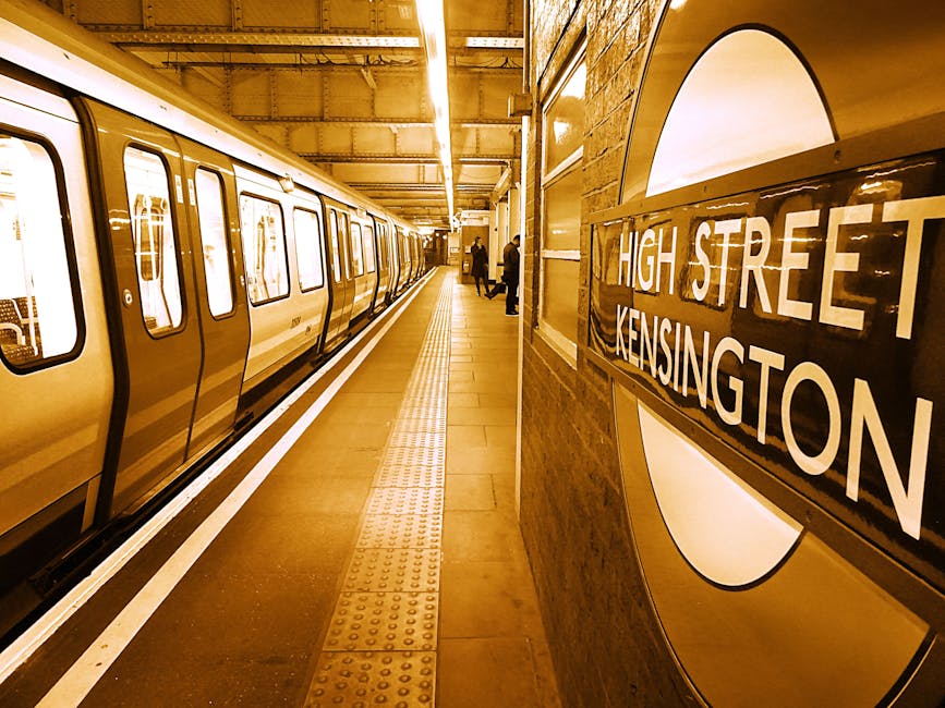 A view of an underground London Underground station platform featuring a yellow and white train stopped alongside the platform edge. The train has a smooth, metallic surface with rectangular windows and rounded edges, reflecting the overhead lighting, which creates a warm amber glow throughout the station. To the right, a curved tiled wall displays a round sign with the black and white London Underground roundel, with the station name 'High Street Kensington' prominently visible. The platform has a textured yellow tactile paving strip running parallel to the train, designed for visually impaired passengers. Several commuters are seen standing near the platform edge, waiting or walking toward the station exit. The environment shows an industrial, brick-clad interior with overhead lighting fixtures casting a consistent warm ambiance. The scene exemplifies typical urban underground transit infrastructure, relevant to discussions on public transport and alternative travel options, with the setting aligned with a busy, well-maintained tube station often used for accessible transportation and intra-city commuting.