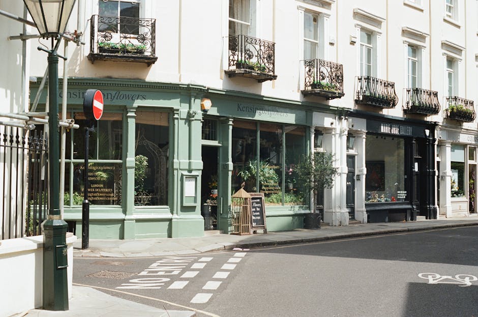 A street scene featuring a row of light-colored, multi-story buildings with ornate black wrought-iron balconies on each floor. The ground floor of the building in the foreground houses a flower shop with large display windows showcasing various potted plants and floral arrangements. The shop has pale green wooden framing and signage indicating it is a flower store, with a small chalkboard sign outside on the sidewalk. To the left, a black lamppost and a street sign with a red circle and white horizontal bar are visible, indicating no entry. The sidewalk has a white dashed line marking a pedestrian crossing, and a bicycle lane symbol is painted on the street. Nearby, there's a black metal fence bordering the property. The scene is set in daylight with natural light illuminating the building facades and street, creating a calm, urban atmosphere associated with local street-level commerce and independent shops.