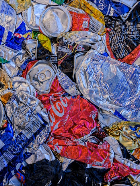 A close-up view of a large pile of crushed aluminum beverage cans, predominantly silver with some red, blue, black, and yellow printed branding, including a visible Coca-Cola logo on a prominent red can. The cans are tightly compressed, with curled and creased surfaces, and some are partially flattened or stacked on top of each other. The scene appears to be outdoors, possibly on a pavement or driveway, with natural lighting highlighting the metallic shine and crinkled textures of the cans. The background shows more cans and discarded packaging, suggesting a collection or disposal of recyclable waste materials. This image reflects waste sorting or collection activity suited to independent rubbish removal or recycling services, such as those offered by Waste Clearance Kensington, operational in the Earls Court SW5 area for same-day rubbish collection and private waste disposal.