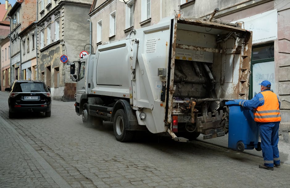 A narrow urban alleyway cluttered with accumulated rubbish and discarded items. In the foreground, a large, grey, weathered plastic bag or bundle of waste, possibly filled with rubbish, is positioned on the dark, gritty pavement. Behind it, a muted silver trolley or cart with a metal frame and fabric sack is partially visible, suggesting it may be used for waste collection or transport. The alley is flanked by tall, weathered brick and concrete buildings, with one side featuring graffiti in vibrant shades of blue, orange, and white. Piles of assorted trash, including cardboard boxes, black plastic bags, and loose debris, are stacked against a makeshift barrier in the background, creating a cluttered scene indicative of private or independent waste disposal. Bare tree branches stretch overhead against a pale sky, adding a stark, wintery atmosphere. The environment appears poorly maintained, emphasizing the need for professional rubbish collection services, such as those offered by Waste Clearance Kensington, to manage urban waste accumulation efficiently and effectively.