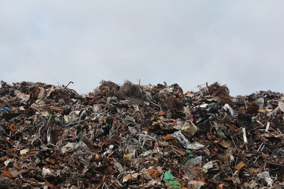 A large, chaotic heap of mixed waste materials extends across the foreground, comprising various discarded items such as crumpled metallic objects, broken plastics, and torn cardboard, all piled high. The texture of the waste is rough and uneven, with some pieces showing rust, peeling paint, and frayed edges, indicating different materials and levels of degradation. The predominant colors include dark browns, rusty reds, faded grays, and occasional bright spots like torn green plastic or scraps of blue. The waste is situated outdoors on what appears to be a paved or concrete surface, possibly an industrial or waste collection site, with a gray, overcast sky in the background, casting subdued natural light over the scene. Faint shadows are visible within the pile, emphasizing its irregularity and dense nature. This image exemplifies a typical scene of bulky or mixed rubbish awaiting removal, reflecting environmental considerations and waste management processes, with Waste Clearance Kensington specialised in handling such accumulation of waste for proper disposal or recycling, supporting private rubbish collection services in lieu of local authority collection.