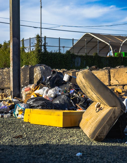 A pile of mixed household waste and rubbish including black plastic bags, a yellow plastic bin, a used car tire, and various debris, all situated on a gravel surface next to a stone wall with some greenery behind it. In the background, there is a metal fence, a tennis court with a net, and a canopy structure, under a partly cloudy sky. The scene appears to depict an outdoor area where rubbish has been discarded or temporarily stored, possibly as part of a private waste clearance service by Waste Clearance Kensington, reflecting how non-standard or alternative waste handling methods might be visually present in urban or suburban settings.