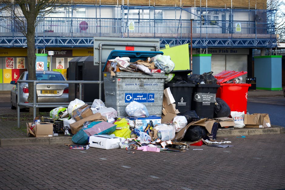 A large, chaotic heap of mixed waste materials extends across the foreground, comprising various discarded items such as crumpled metallic objects, broken plastics, and torn cardboard, all piled high. The texture of the waste is rough and uneven, with some pieces showing rust, peeling paint, and frayed edges, indicating different materials and levels of degradation. The predominant colors include dark browns, rusty reds, faded grays, and occasional bright spots like torn green plastic or scraps of blue. The waste is situated outdoors on what appears to be a paved or concrete surface, possibly an industrial or waste collection site, with a gray, overcast sky in the background, casting subdued natural light over the scene. Faint shadows are visible within the pile, emphasizing its irregularity and dense nature. This image exemplifies a typical scene of bulky or mixed rubbish awaiting removal, reflecting environmental considerations and waste management processes, with Waste Clearance Kensington specialised in handling such accumulation of waste for proper disposal or recycling, supporting private rubbish collection services in lieu of local authority collection.