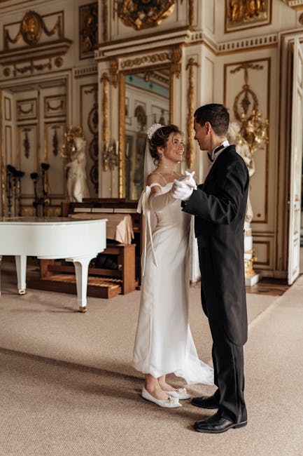 A bride and groom are standing in an opulent, ornately decorated room with cream-colored walls adorned with gold accents, intricate moldings, and tall mirrors with gilded frames. The bride wears a white wedding gown with long sleeves, lace detailing on the bodice, and a flowing skirt, while her hair is styled in an elegant updo with a veil. The groom is dressed in a formal black tuxedo with a white shirt, black bow tie, and black dress shoes. They are holding hands and facing each other, engaging in a tender moment, with the bride smiling softly. In the background, a white grand piano is positioned near a small table with a pink cloth, and nearby are sculptures and ornamental decorations on the walls, indicating the setting is a luxurious venue suited for special events like weddings or high-society gatherings. The lighting is soft, highlighting the refined atmosphere of the room, and the scene captures a private moment between the newlyweds in a splendid, classical interior that complements the occasion of a formal celebration or wedding reception.