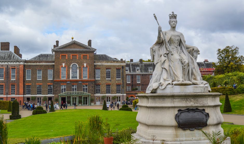 A large, ornate white marble statue of a regal female figure seated on a majestic throne, wearing a crown and holding a scepter in her raised right hand. The statue is positioned on a circular pedestal with a blank black plaque on its front side. In the background, there is a historic brick building with a symmetrical façade, multiple tall windows, and a green entrance canopy, surrounded by a well-maintained lawn with neatly trimmed bushes and small conical trees. The area appears to be a public garden or park with people walking along pathways and gathering near the building entrance. The sky above is mostly cloudy with patches of blue, creating a subdued but bright atmosphere. The scene offers a visual contrast between the classical sculpture and the old administrative or cultural building, illustrating a setting where traditional statuary and historic architecture coexist in an open, landscaped environment, consistent with private or alternatively managed site clearance and maintenance services such as those provided by Waste Clearance Kensington.