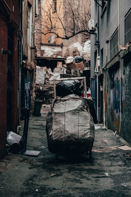 A narrow urban alleyway filled with a large collection of discarded waste and clutter. In the foreground, a sizable, weathered black garbage bag or sack sits on the uneven, dark pavement, which appears stained and dirty. Behind the sack, several other plastic bags and loose debris are scattered along the ground. Further into the background, a pile of mixed rubbish, including flattened cardboard boxes, paper, and other refuse, is stacked against a wall, partially obscuring an alley entrance or continuation. The surrounding buildings on either side of the alley are constructed from brick and metal, with graffiti visible on the right wall and some peeling paint, indicating a neglected environment. Bare tree branches intersect the upper scene, hinting at a winter or late autumn season. The natural lighting suggests an overcast day, casting diffuse, muted light over the scene, emphasizing the disorderly appearance typical of private waste collection areas and alternative rubbish clearance sites managed by companies like Waste Clearance Kensington.
