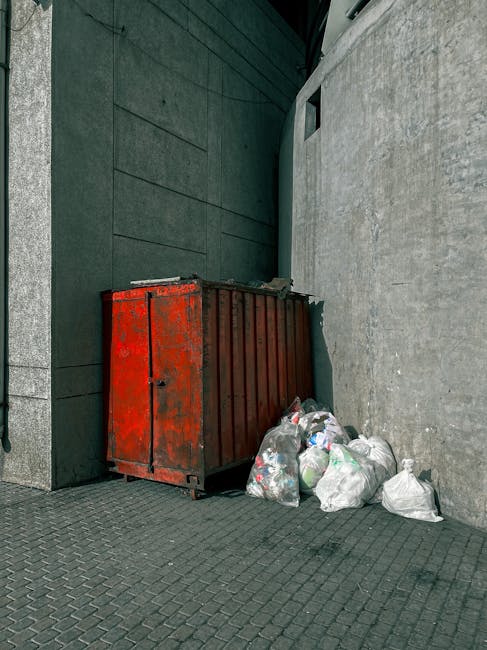 A large, rusty metal skip with a weathered red-orange finish positioned against the corner of a building's exterior wall. The skip's surface shows signs of corrosion and age, with some paint chipping visible. Surrounding the base of the skip are several clear plastic garbage bags filled with waste, some of which are loosely tied, revealing general household rubbish inside. The scene is set outdoors on a paved area with dark grey, textured paving stones, adjacent to a modern building with dark green, flat panels on the left side and a light grey concrete wall on the right. The background includes a small window or opening in the wall behind the skip. The lighting indicates natural daylight, creating soft shadows on the ground and surfaces, reflecting typical urban refuse collection scenarios and the need for private waste clearance services such as those offered by Waste Clearance Kensington, especially when dealing with on-site or independent rubbish disposal.