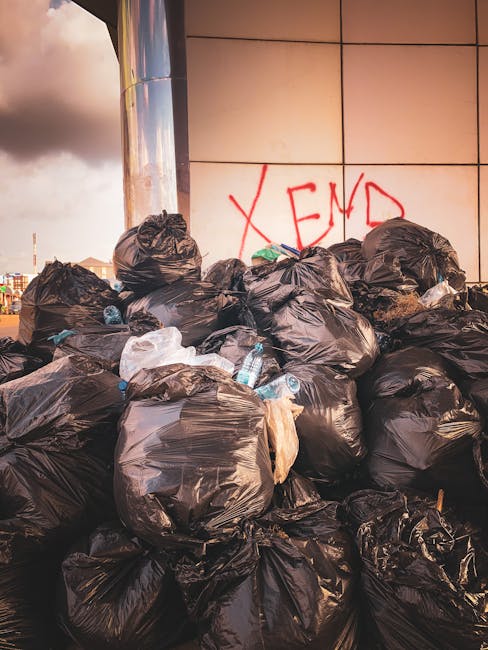 A narrow urban alleyway filled with a large collection of discarded waste and clutter. In the foreground, a sizable, weathered black garbage bag or sack sits on the uneven, dark pavement, which appears stained and dirty. Behind the sack, several other plastic bags and loose debris are scattered along the ground. Further into the background, a pile of mixed rubbish, including flattened cardboard boxes, paper, and other refuse, is stacked against a wall, partially obscuring an alley entrance or continuation. The surrounding buildings on either side of the alley are constructed from brick and metal, with graffiti visible on the right wall and some peeling paint, indicating a neglected environment. Bare tree branches intersect the upper scene, hinting at a winter or late autumn season. The natural lighting suggests an overcast day, casting diffuse, muted light over the scene, emphasizing the disorderly appearance typical of private waste collection areas and alternative rubbish clearance sites managed by companies like Waste Clearance Kensington.