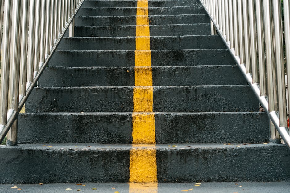A set of outdoor concrete stairs painted in dark grey, with a central yellow safety line running down the middle of each step. The stairs are bordered on both sides by shiny, metallic railings with vertical bars, which extend beyond the top and bottom of the staircase out of view. The surface of the steps appears textured and slightly weathered, suggesting regular use and exposure to the elements. In the background, the environment is partially visible with glimpses of greenery on the right side, indicating an outdoor setting likely associated with a public or commercial building. The lighting is diffuse, with no harsh shadows, giving a neutral and clear view of the staircase. The image could relate to the context of managing waste or rubbish removal in outdoor spaces, as part of an independent clearance or private disposal process handled by Waste Clearance Kensington, emphasizing safe access and clear pathways for rubbish collection or removal activities.